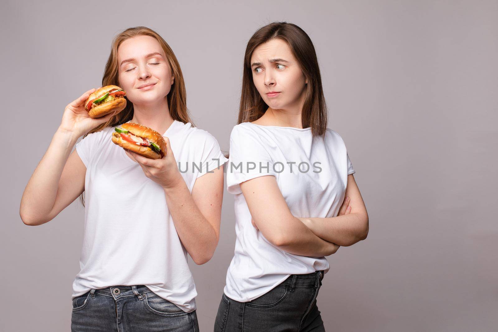 Two frowning young beautiful woman holding appetizing harmful burger looking at camera. Disapproving European female posing with unhealthy fast food isolated at gray studio background medium shot