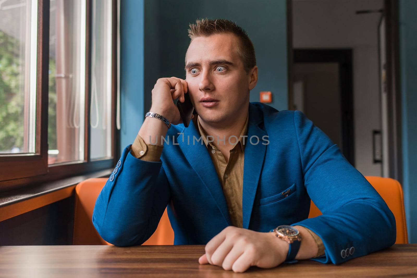 An adult stylish, businessman of European appearance portrait sits in a jacket at a table in a cafe and talks on a smartphone or mobile phone and looking out the window.