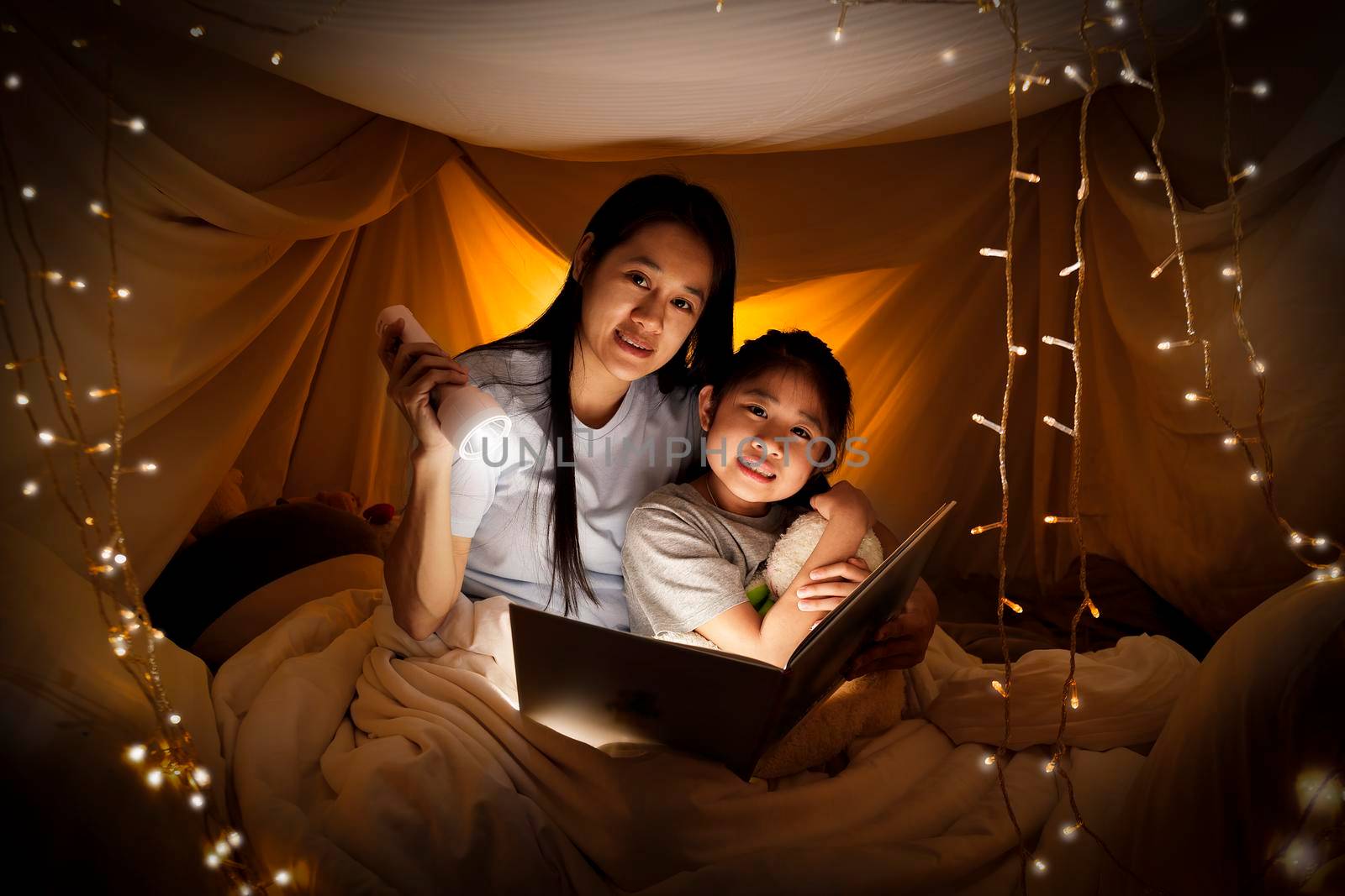 Family concept. Mother and child daughter reading book with flashlight together in children tent before bedtime. Happy mother read story book to her daughter in bed sheet tent
