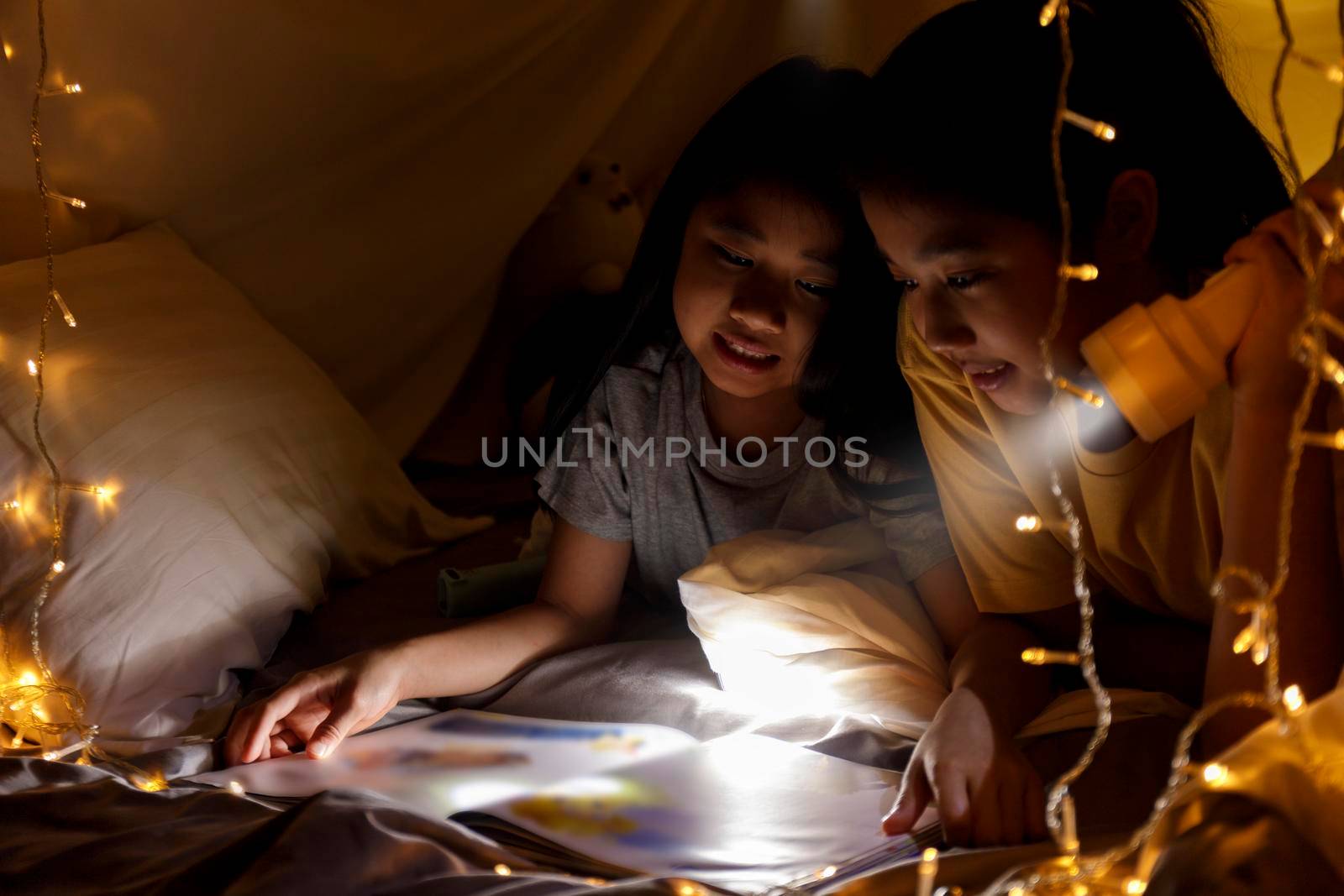 Family concept. elder sister and sister reading book with flashlight together before bedtime. Sister read story book together in bed sheet tent. focus selective little sister. With film grain effect