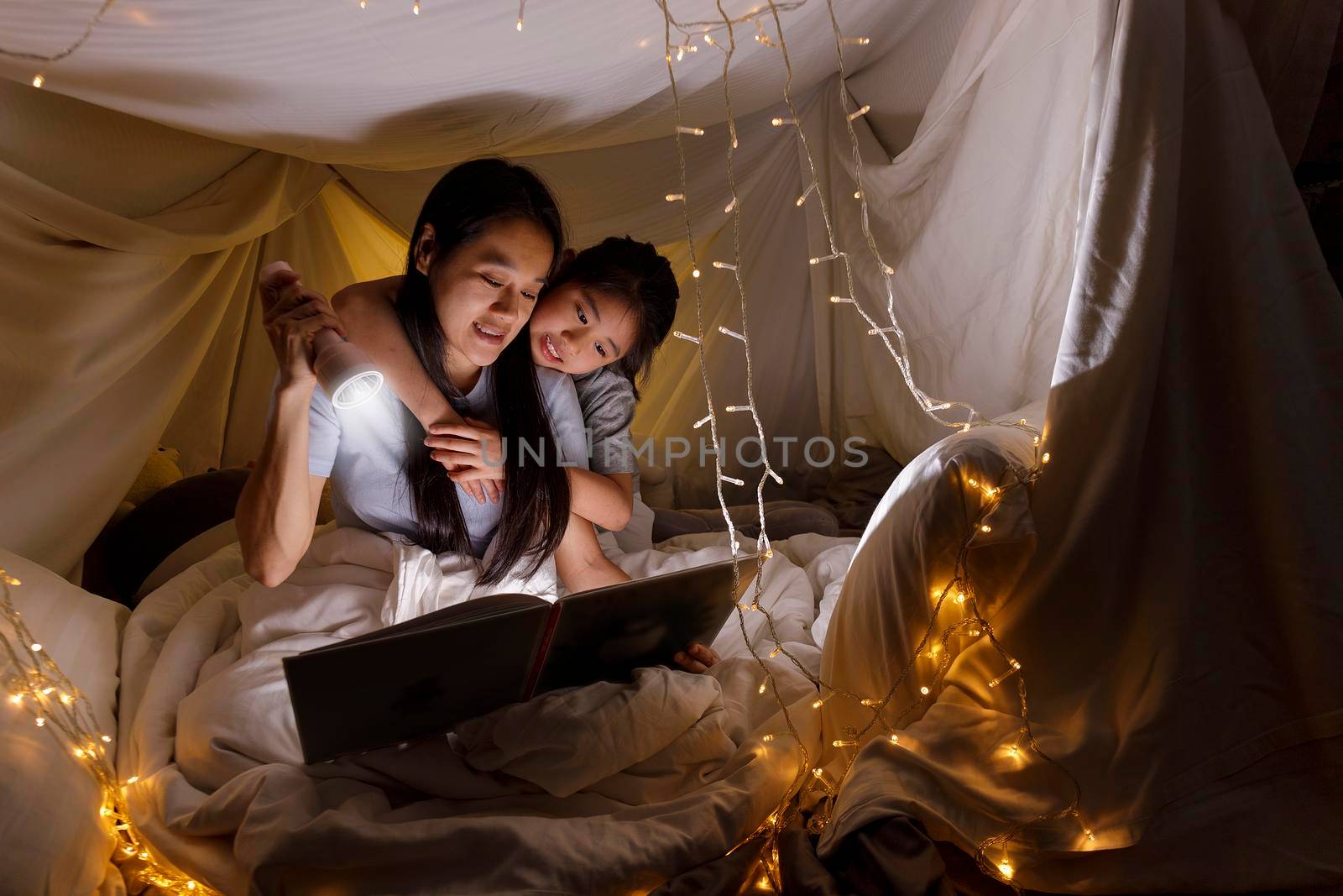 Family concept. Mother and child daughter reading book with flashlight together in children tent before bedtime. Happy mother read story book to her daughter in bed sheet tent
