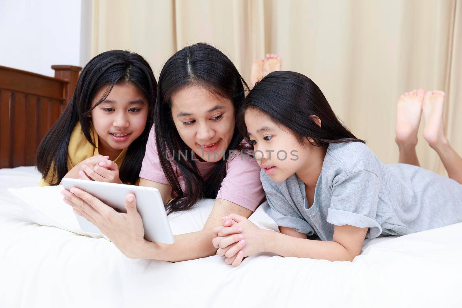 Happy Asian family enjoy and relax on bed in bedroom. mother and daughters enjoy using tablet together on bed.  Family concept. by Satrinekarn