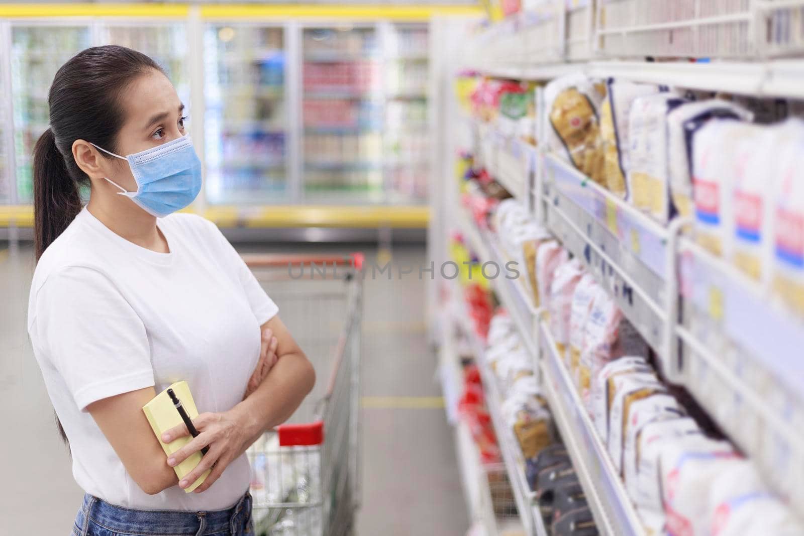 Asian woman wear face mask push shopping cart in supermarket. female looking grocery to buy something During coronavirus crisis or covid19 outbreak. Women wearing protective face mask new normal concept by Satrinekarn