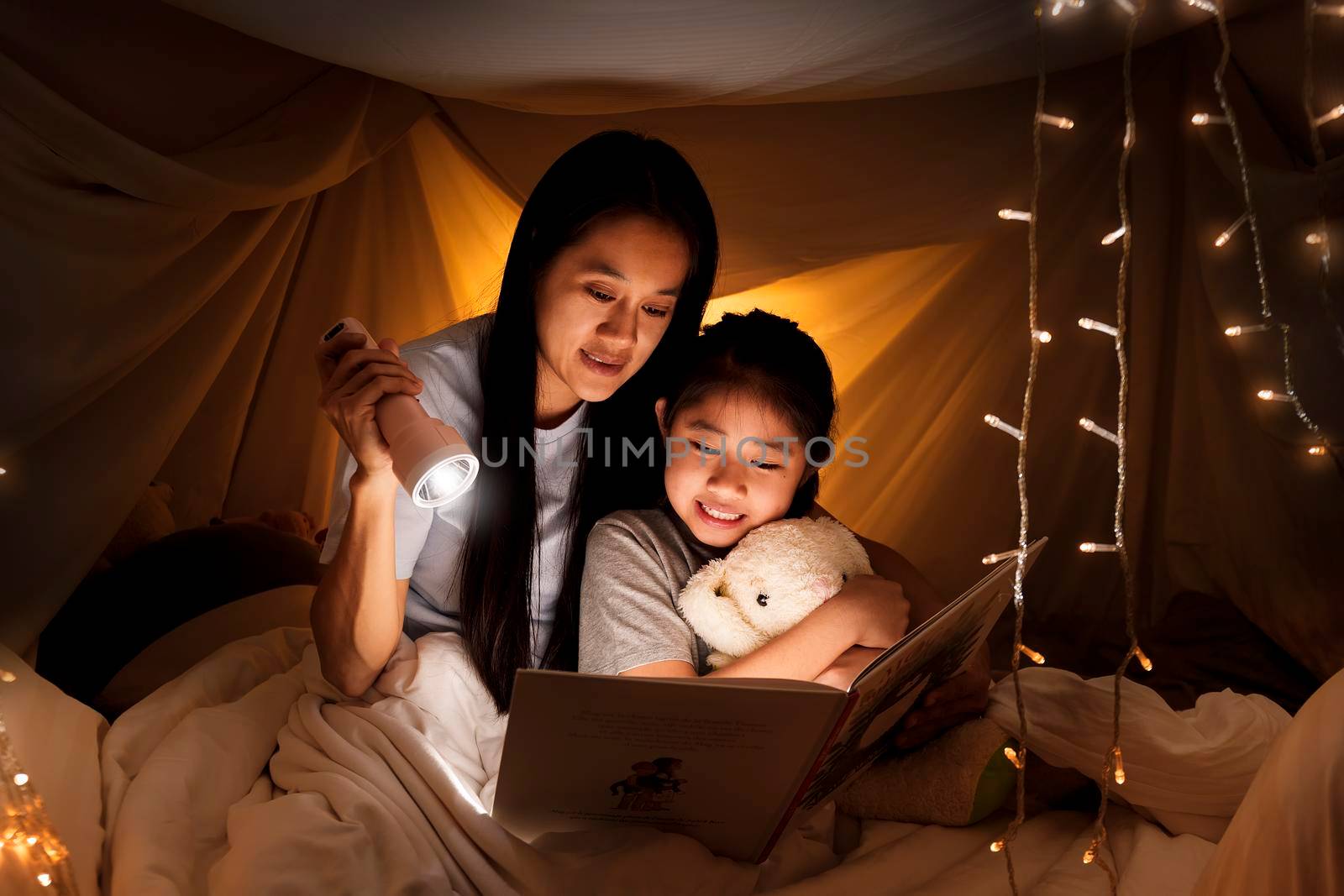 Family concept. elder sister and sister reading book with flashlight together before bedtime. Sister read story book together in bed sheet tent. focus selective little sister. With film grain effect by Satrinekarn