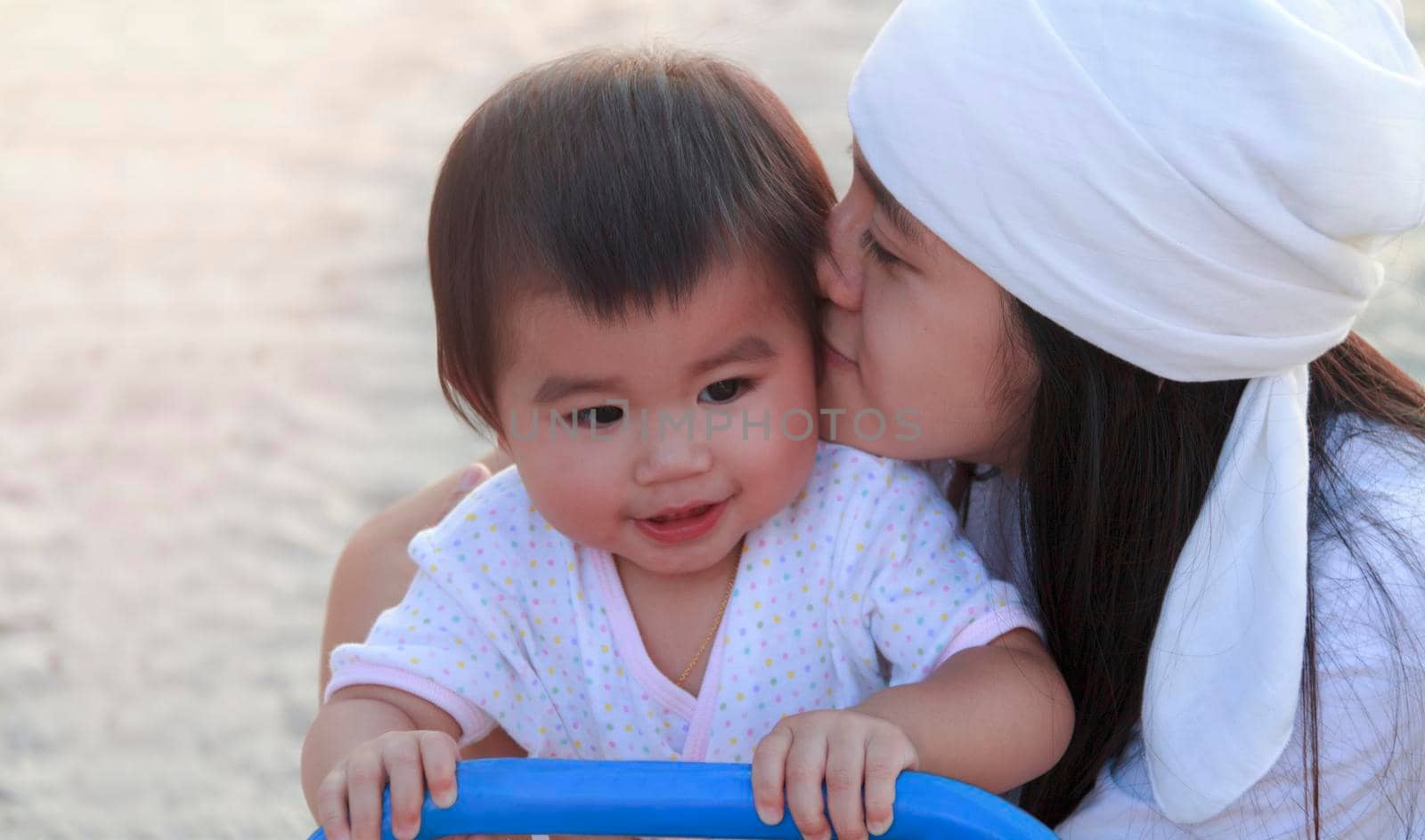 Close up portrait beautiful young asian mother kissing her little baby in park. Happy lovely mother kiss her little daughter. Smiling little girl play at playground with her mother. by Satrinekarn