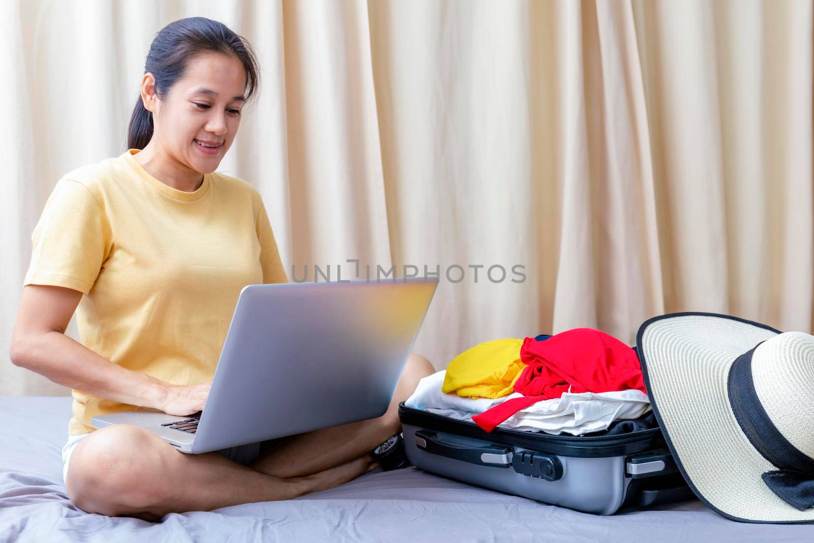 Asian happy woman plan for trip with her friend by laptop on the bed at home. Women with travel bag use notebook computer search for booking hotel frome internet. Holiday summer vacation concept