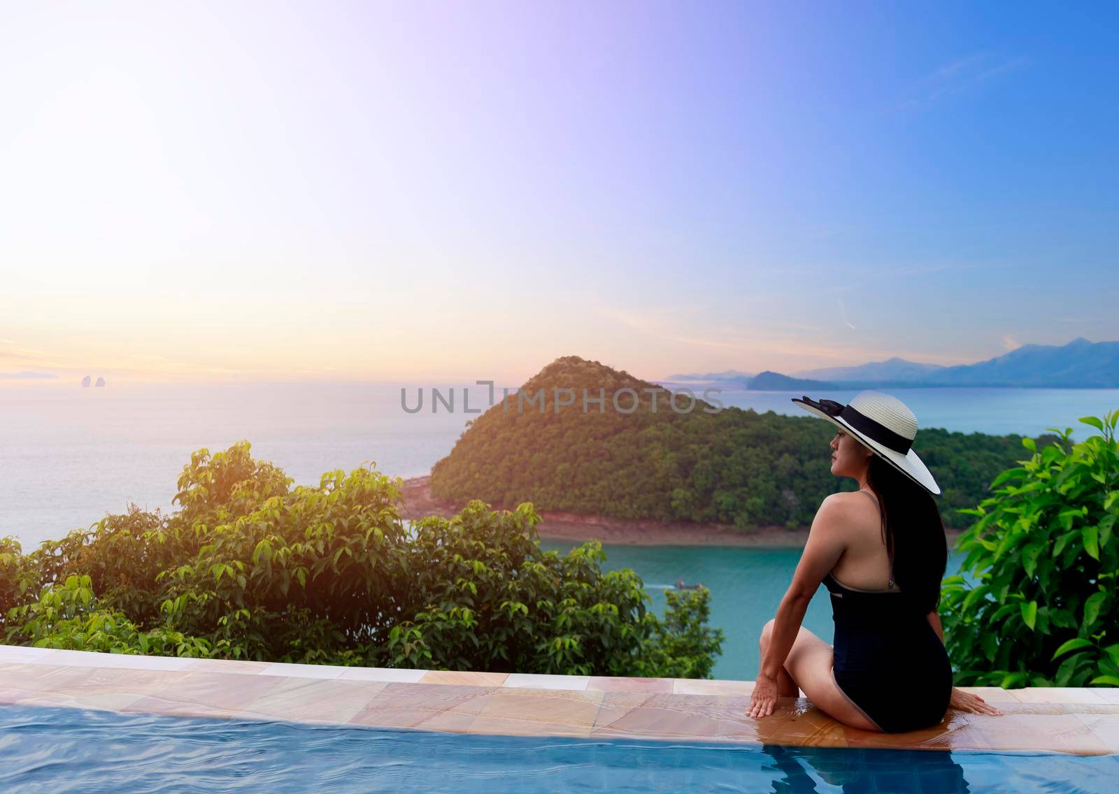 Asian women in black swimsuit and hat siting on side of infinity pool relaxing at sunset time  with sea view during vacation retreat. Women happy and enjoy with holiday summer travel time.