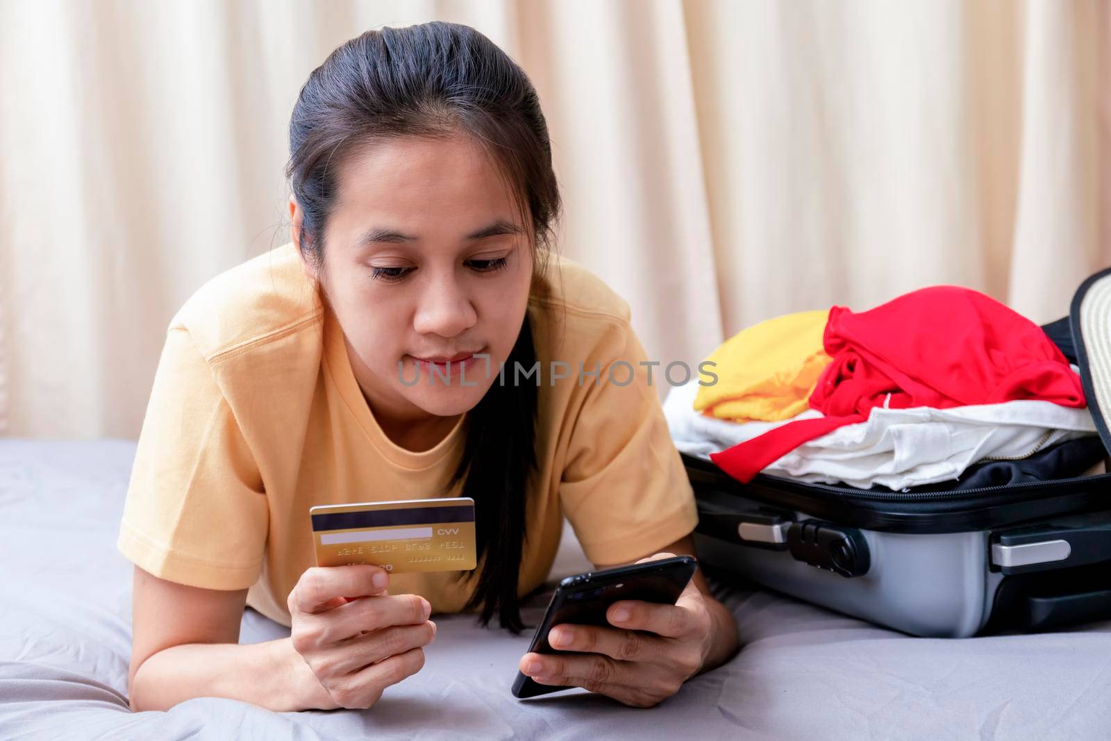 Asian happy woman plan for trip by mobile phone and credit card on the bed at home. Women with travel bag use smart phone and credit card pay booking hotel. Holiday summer concept