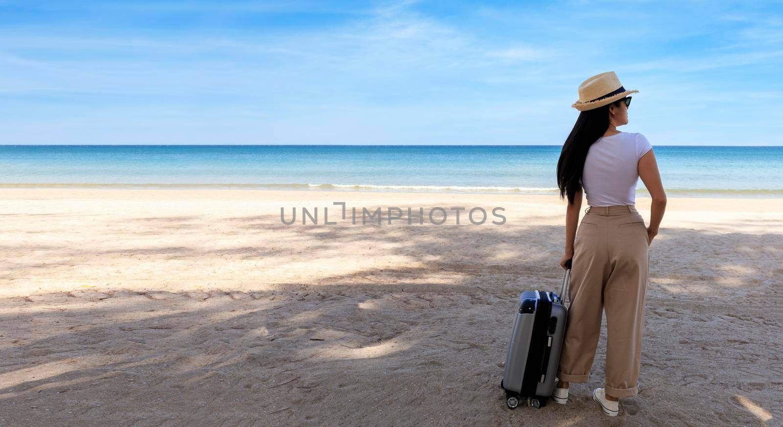 Beautiful young woman wear T-shirt, long pants and straw hat with a suitcase on a tropical beach. Blue sea in the background. Happy travel women standing with travel bag relax on beach. Holiday travel vacation summer