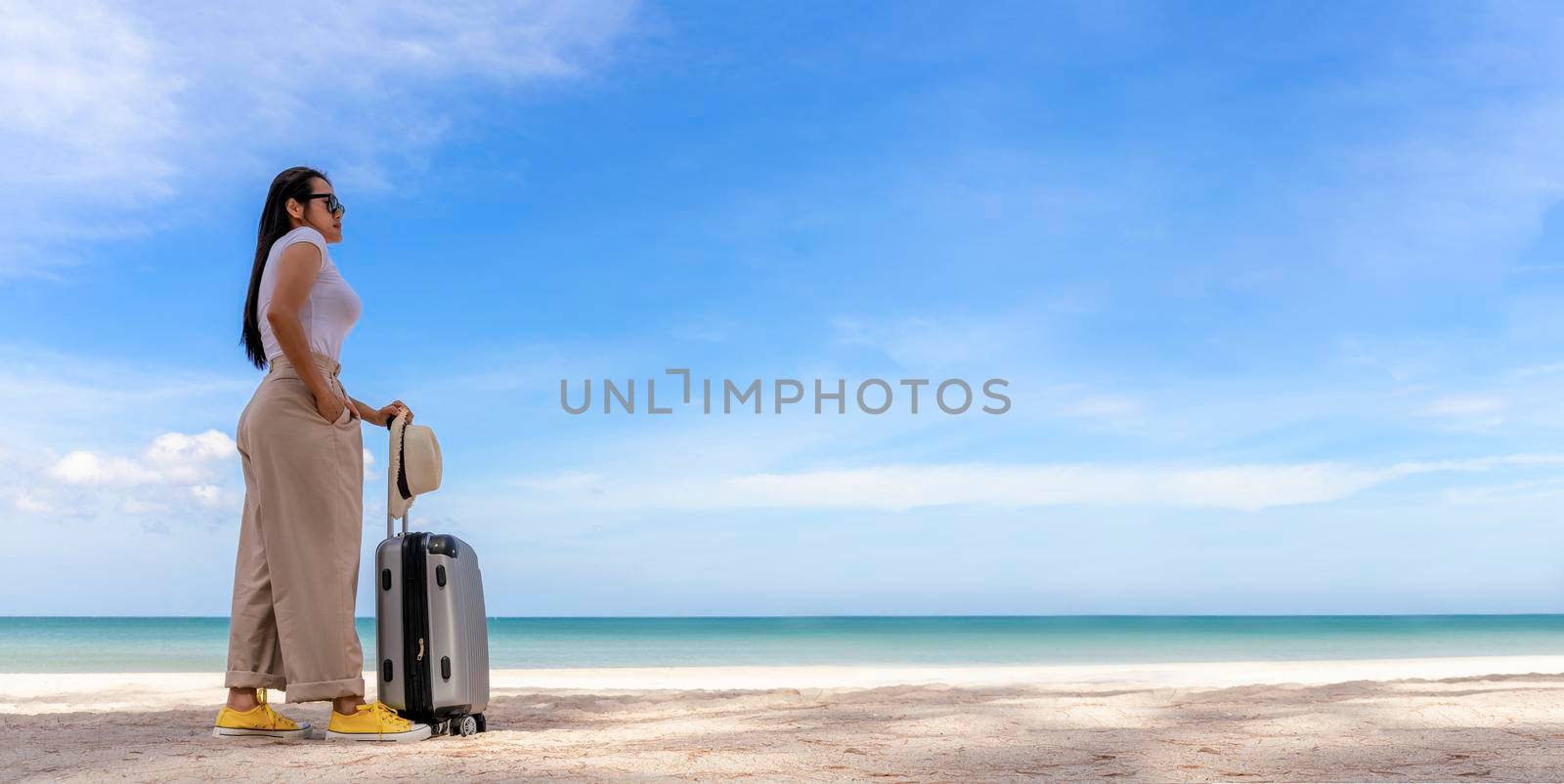 Beautiful young woman wear T-shirt, long pants and straw hat with a suitcase on a tropical beach. Blue sea in the background. Happy travel women standing with travel bag relax on beach. Holiday travel vacation summer