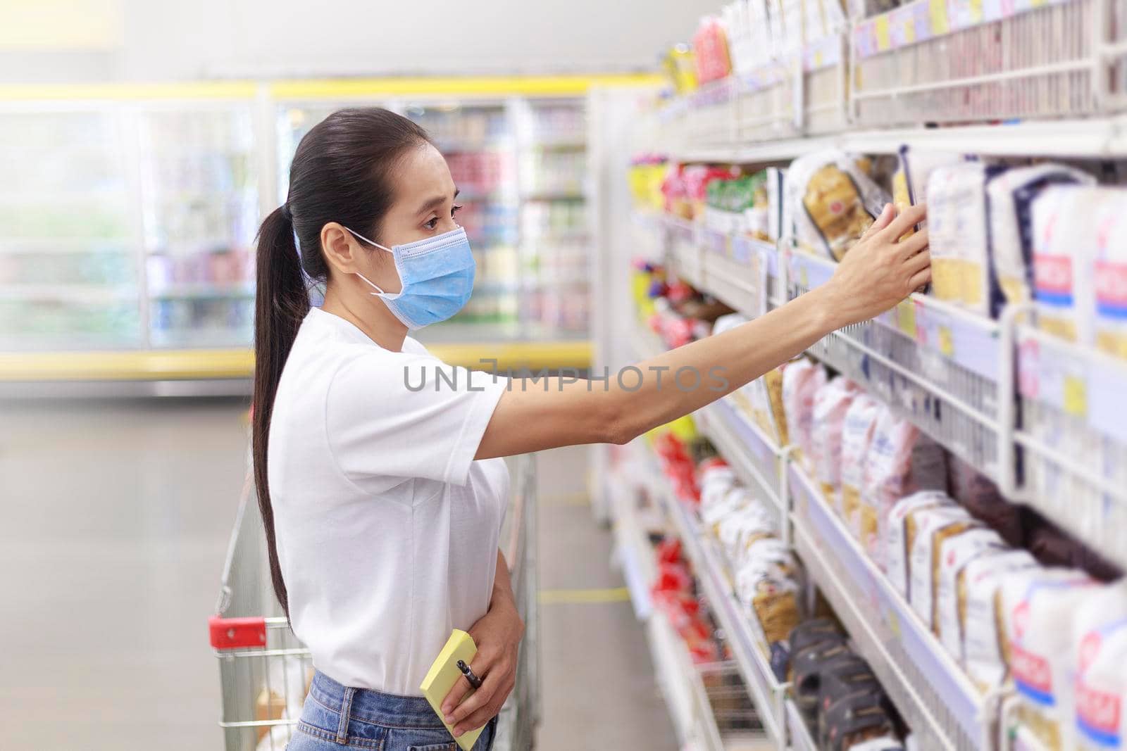 Asian woman wear face mask push shopping cart in supermarket. female looking grocery to buy something During coronavirus crisis or covid19 outbreak. Women wearing protective face mask new normal concept
