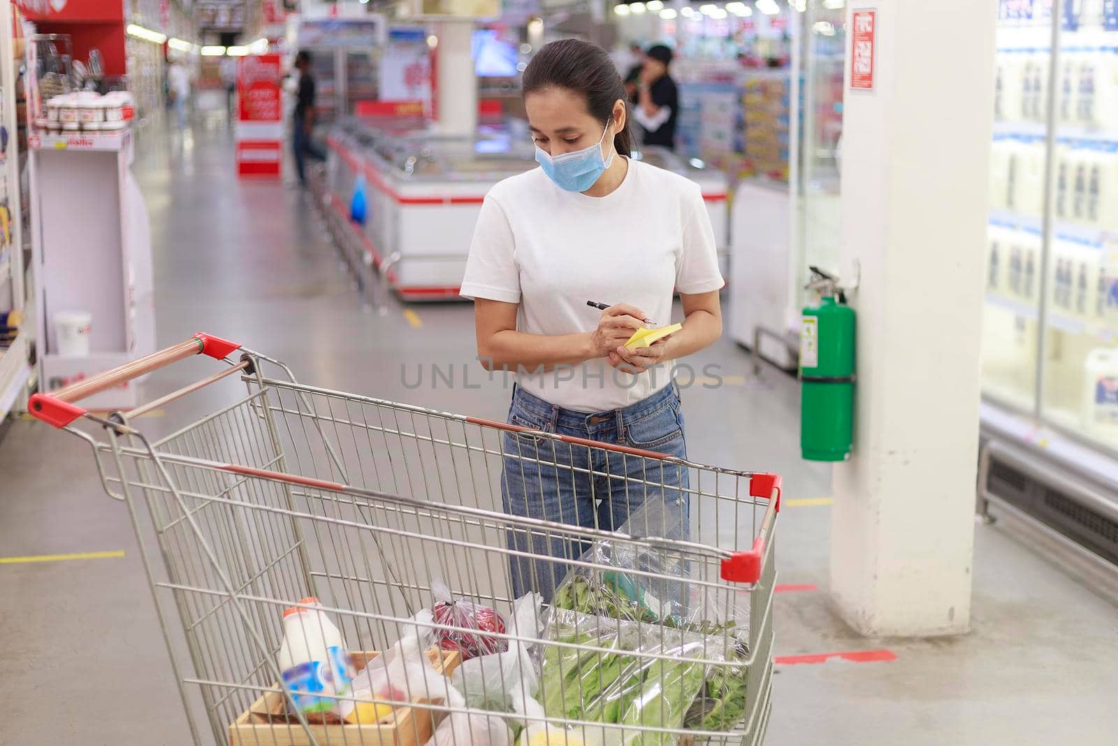 Asian woman wear face mask push shopping cart in supermarket. Girl looking grocery to buy something During coronavirus crisis or covid19 outbreak. Women checking her shopping items on list new normal concept