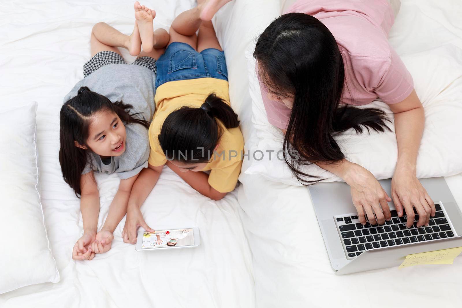 Asian mother and daugthers looking laptop and tablet on bed in bedroom. Business women woking with laptop at home. Girl enjoy speak with father video call  on bed.