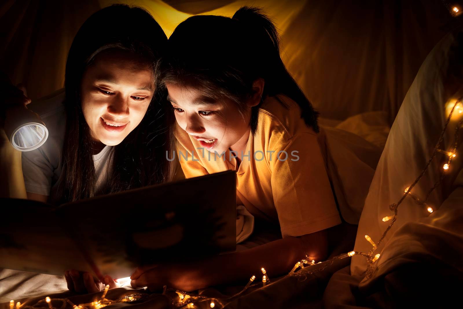 Family concept. Mother and child daughter reading book with flashlight together in children tent before bedtime. Happy mother read story book to her daughter in bed sheet tent
