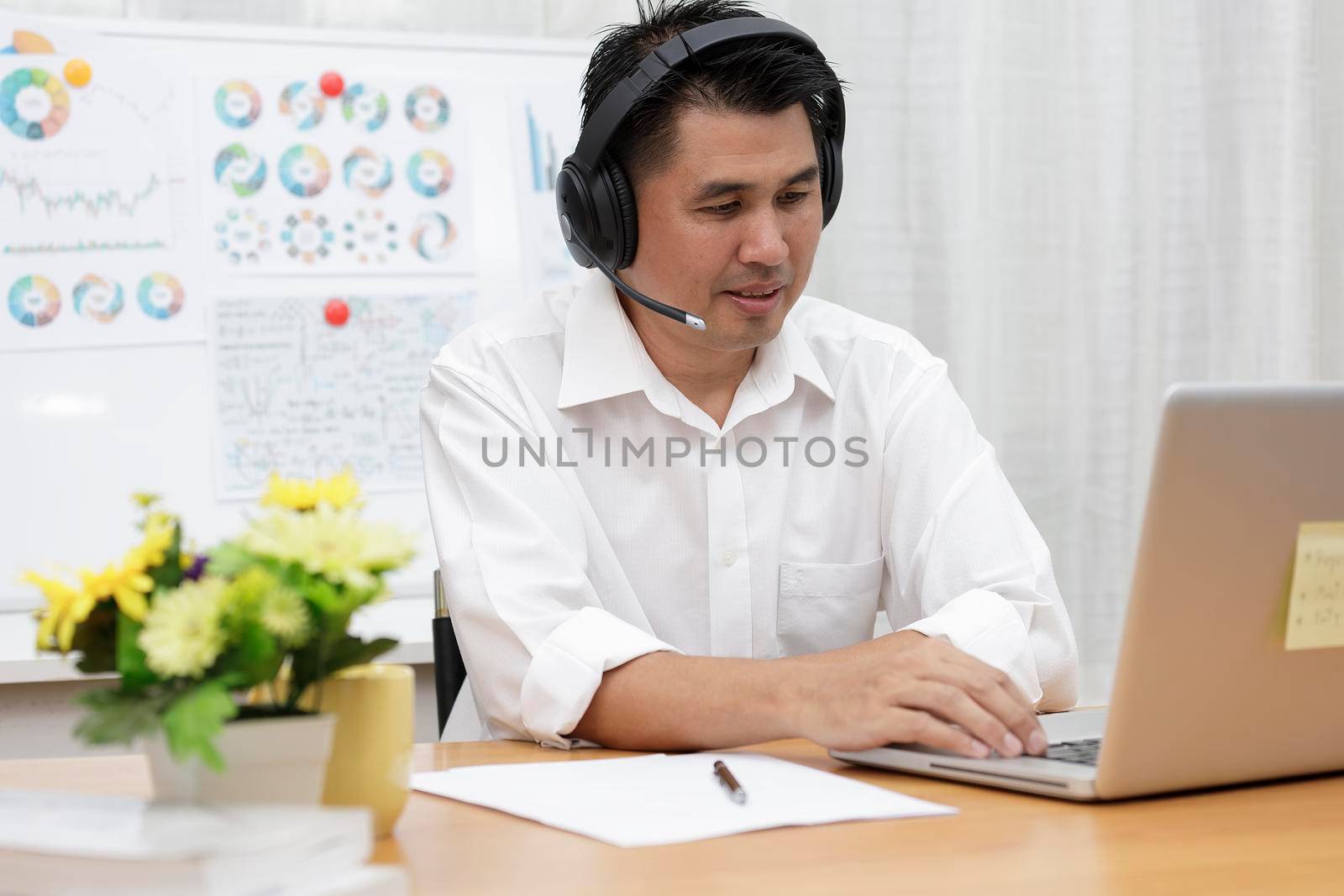 Asian businessman talking with team via video conference writing note on paper. business people using laptop and headset for online meeting. Man using headphone speak with customers. work from home concept.