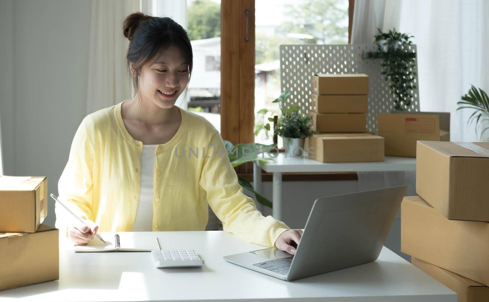 A portrait of Asian woman, e-commerce employee sitting in the office full of packages on the table using a laptop and calculator, for SME business, e-commerce, technology and delivery business..