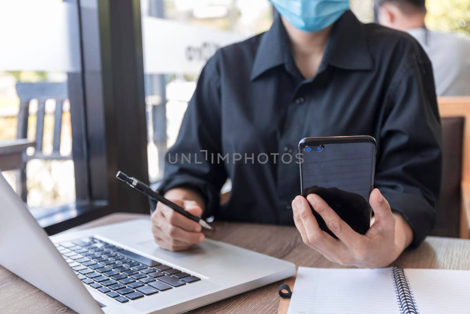 Young woman with protective face mask sitting in coffee shop at wood table using smartphone.and laptop. Girl browsing internet, chatting, blogging. Female holding phone and looking on screen