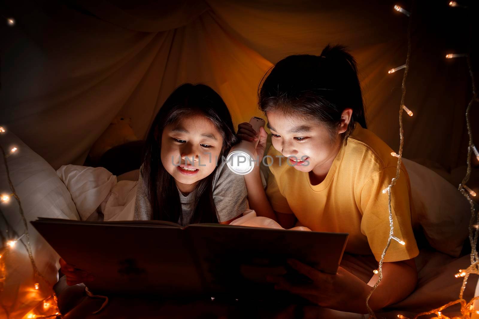 Family concept. elder sister and sister reading book with flashlight together before bedtime. Sister read story book together in bed sheet tent. focus selective little sister. With film grain effect