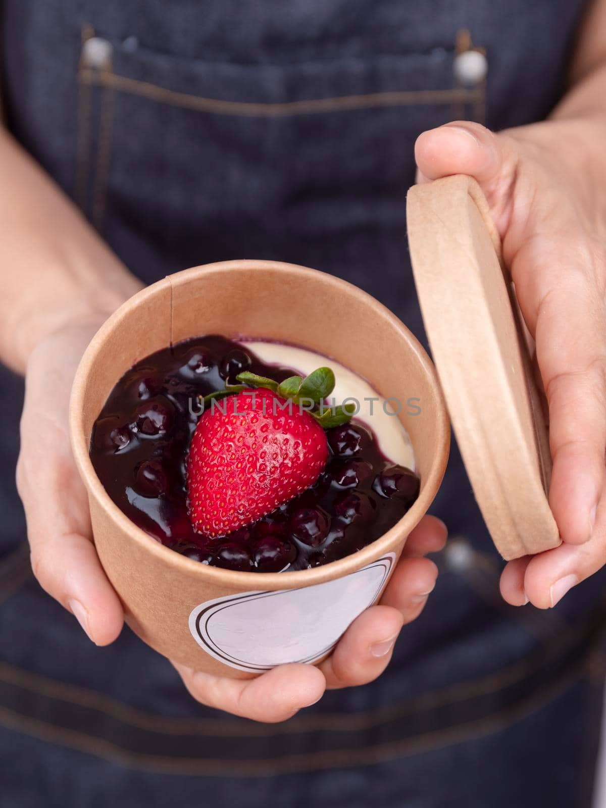 Hands of pastry chef hold blueberry and fresh strawberry cheese pie in paper craft cup with empty label. Homemade blueberry cheese cake in paper cup.