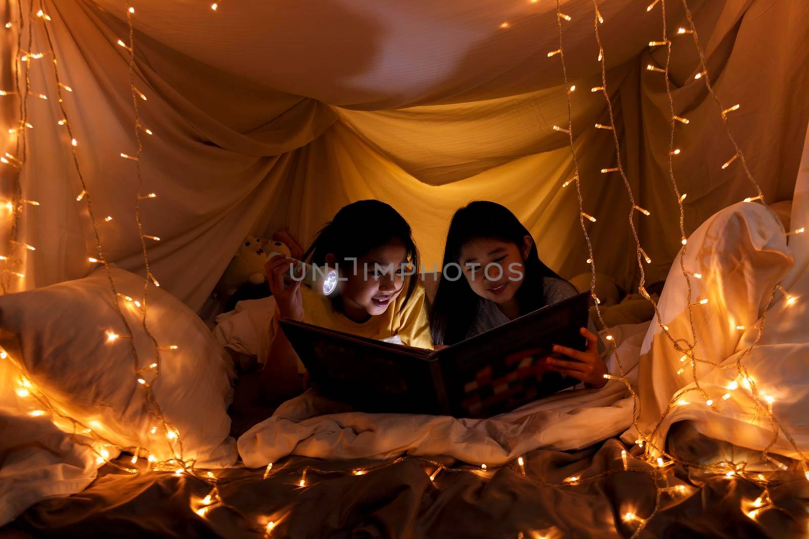 Family concept. elder sister and sister reading book with flashlight together in children tent before bedtime. Happy elder sister read story book to her sister in bed sheet tent by Satrinekarn