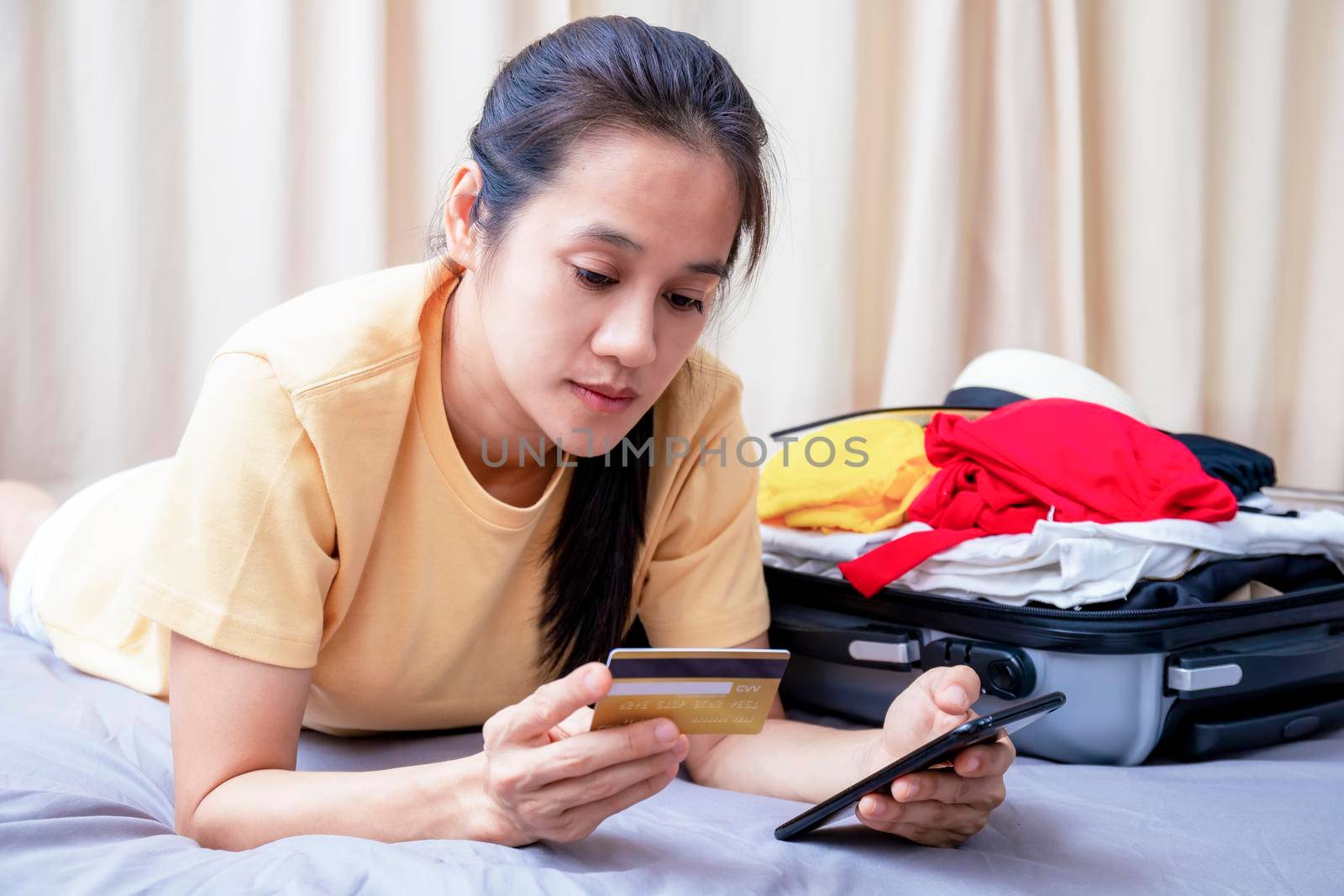Asian happy woman plan for trip by mobile phone and credit card on the bed at home. Women with travel bag use smart phone and credit card pay booking hotel. Holiday summer concept