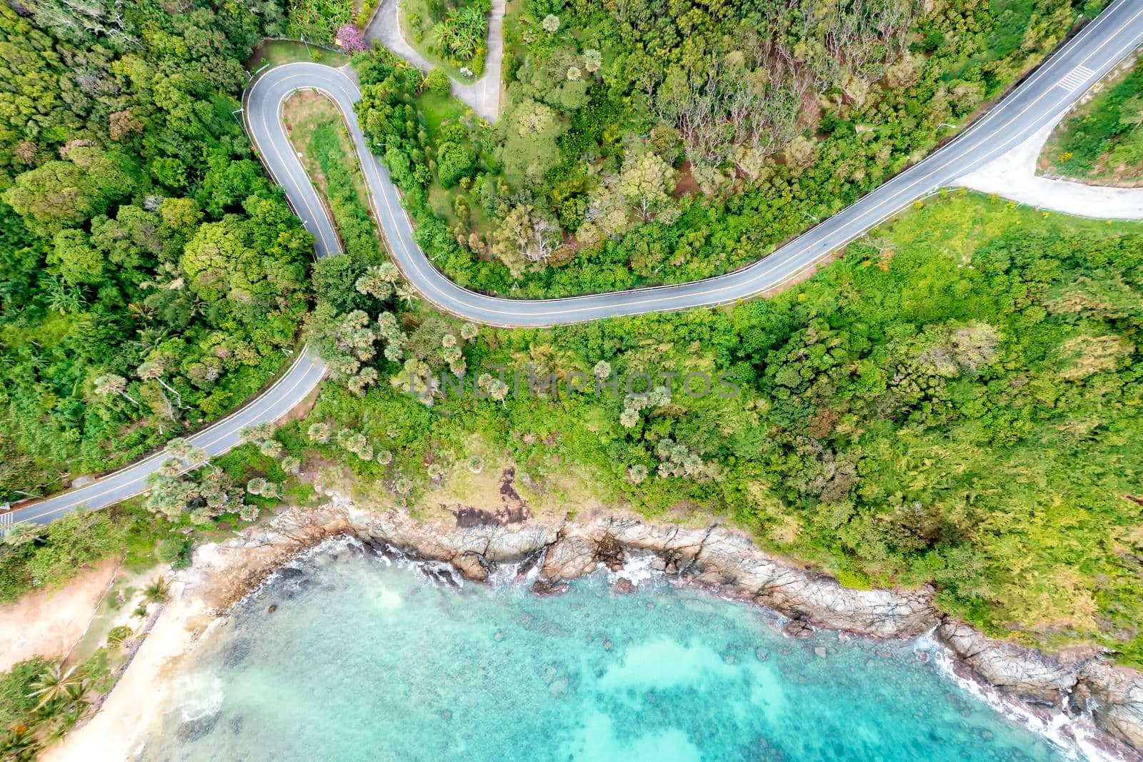 Aerial drone top down view of waves breaking in the sand with road and forest. High angle view of tropical sea with Seafront road. Phuket Thailand curves of coastal road. Phuket is famous tourist destination. Top view traffic road around the beach