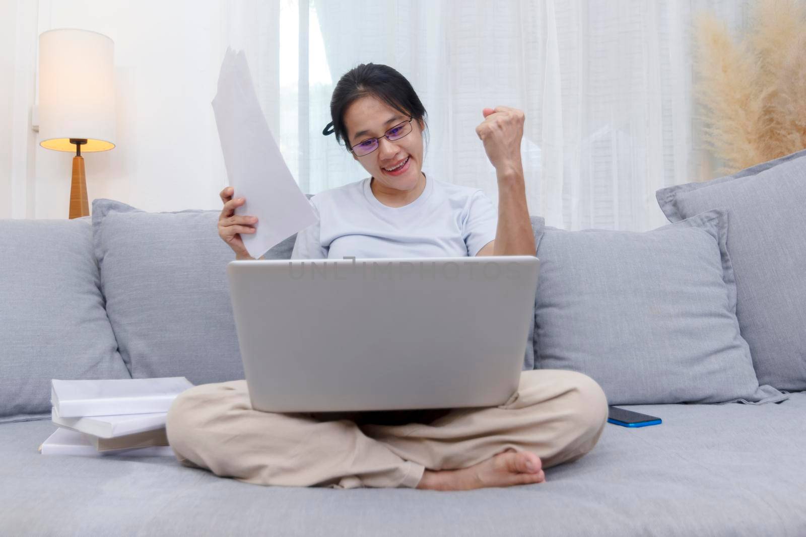 Asian woman celebrating online win success looking laptop at home. Work from home business women sitting on sofa in living room working with laptop online. Young women excited and glad of success with laptop on sofa 