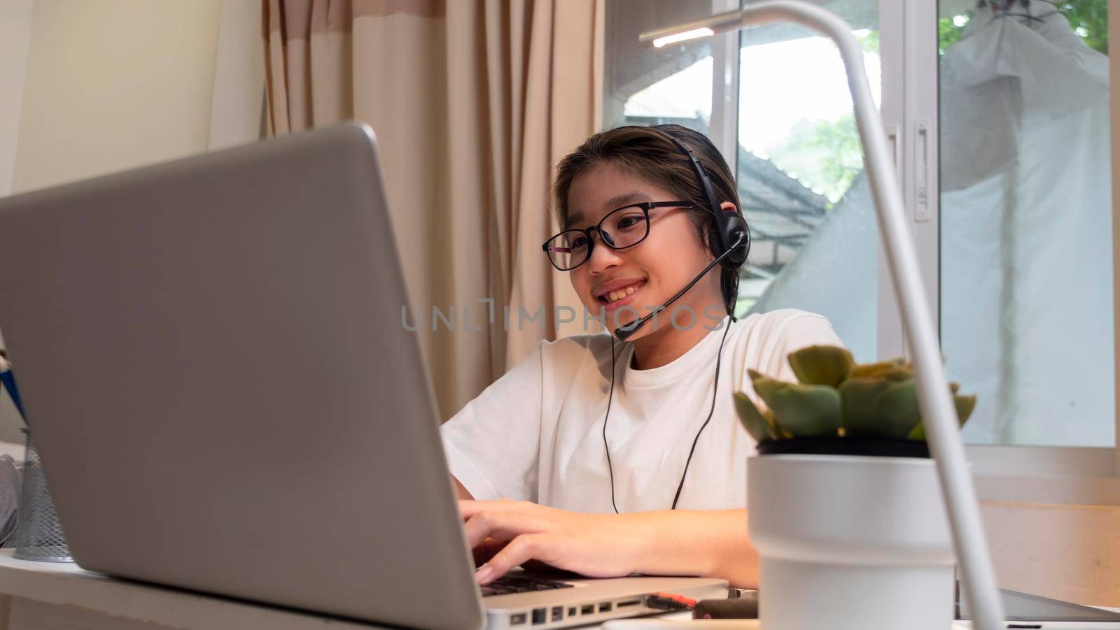  Asian girl in headphones sit at desk study online on laptop. Kid wear headset handwriting in notebook learning using internet lessons on quarantine. Girl student learning virtual internet online class from school due to covid pandemic.