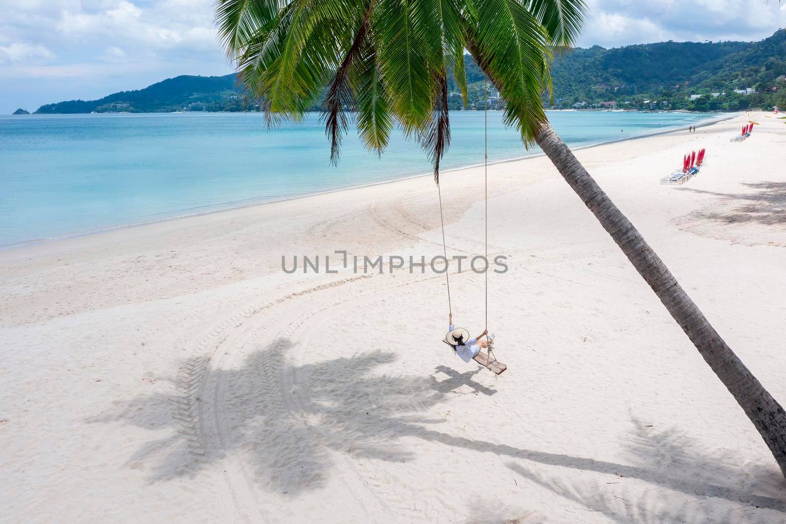 Phuket, Thailand. Tropical beach paradise with beach swing with girl in white shirt. Women relax on swing under coconut palm tree at beautiful tropical beach White sand. Luxury beach scene vacation summer holiday concept.