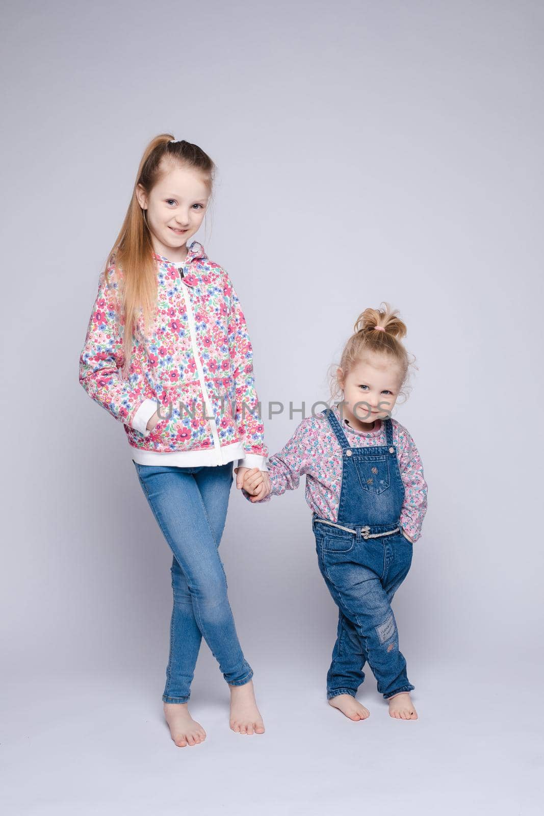 Front view of two pretty sisters keeping hands, looking at camera and smiling on isolated background in studio. Happy kids wearing stylish outfits posing together. Concept of friendship and fashion.