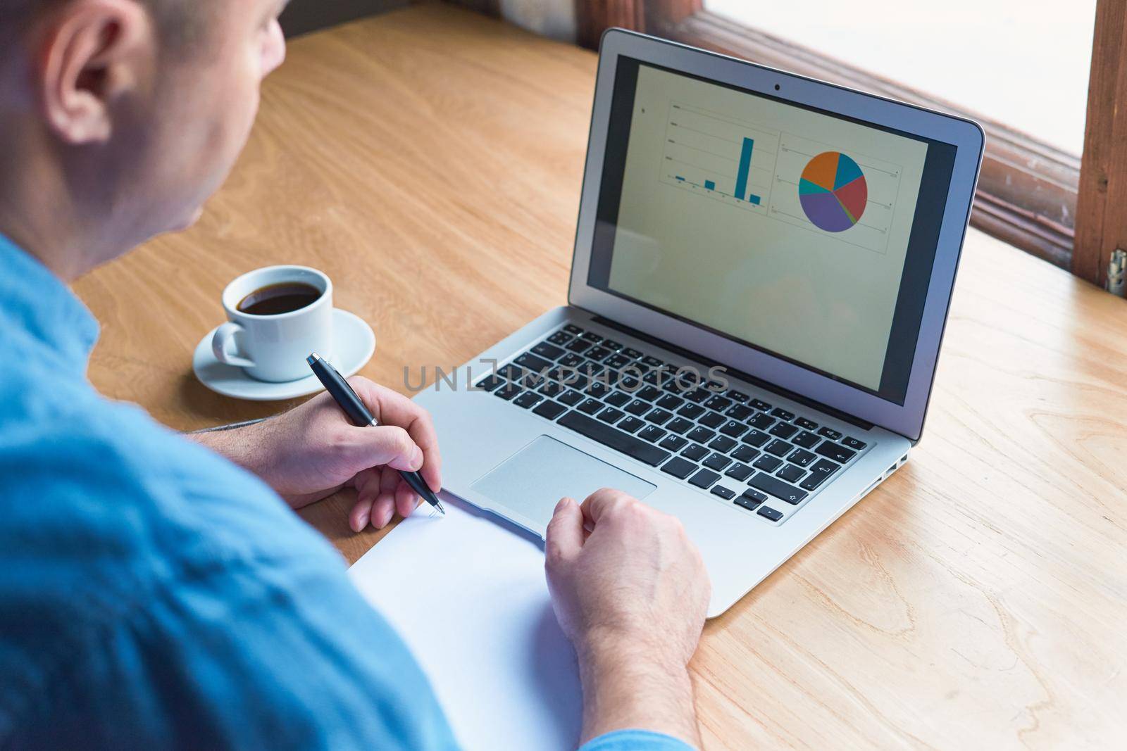 Unrecognizable man writes a plan on paper and looks at charts on a computer screen, laptop. A man with casual clothes in a blue shirt sits in an office in front of a window