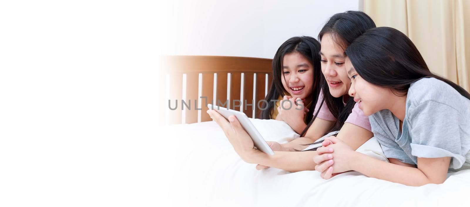 Happy Asian family enjoy and relax on bed in bedroom. mother and daughters enjoy using tablet together on bed.  Family concept. With copy space banner.