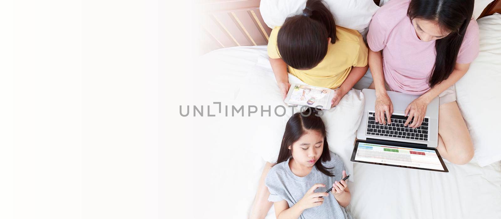 Top view Asian mother and daugthers looking laptop, smart phone and tablet on bed in bedroom. Business women woking with laptop at home. Girl enjoy speak with father video call  on bed. With copy space banner