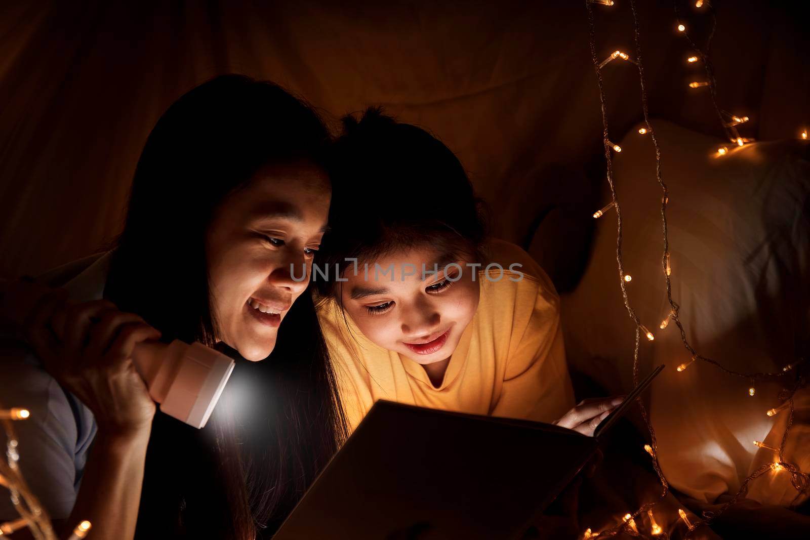 Family concept. elder sister and sister reading book with flashlight together before bedtime. Sister read story book together in bed sheet tent. focus selective little sister. With film grain effect