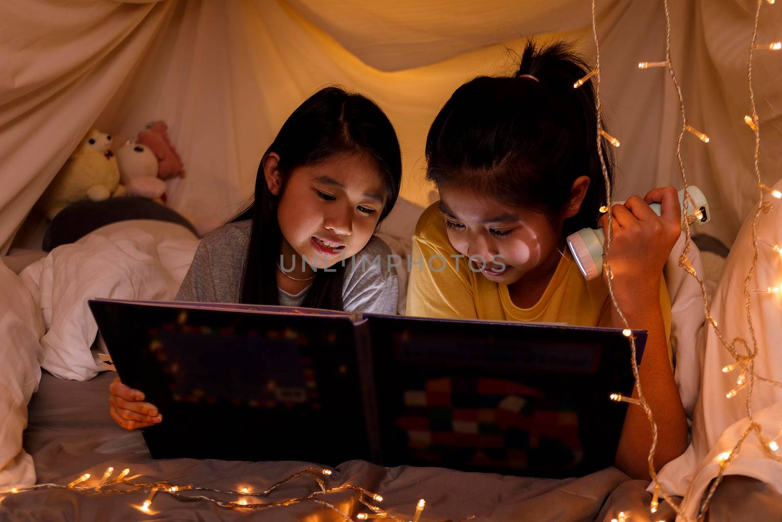 Family concept. elder sister and sister reading book with flashlight together in children tent before bedtime. Happy elder sister read story book to her sister in bed sheet tent
