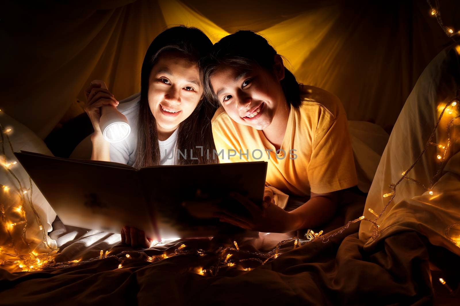 Family concept. Mother and child daughter reading book with flashlight together in children tent before bedtime. Happy mother read story book to her daughter in bed sheet tent
 by Satrinekarn