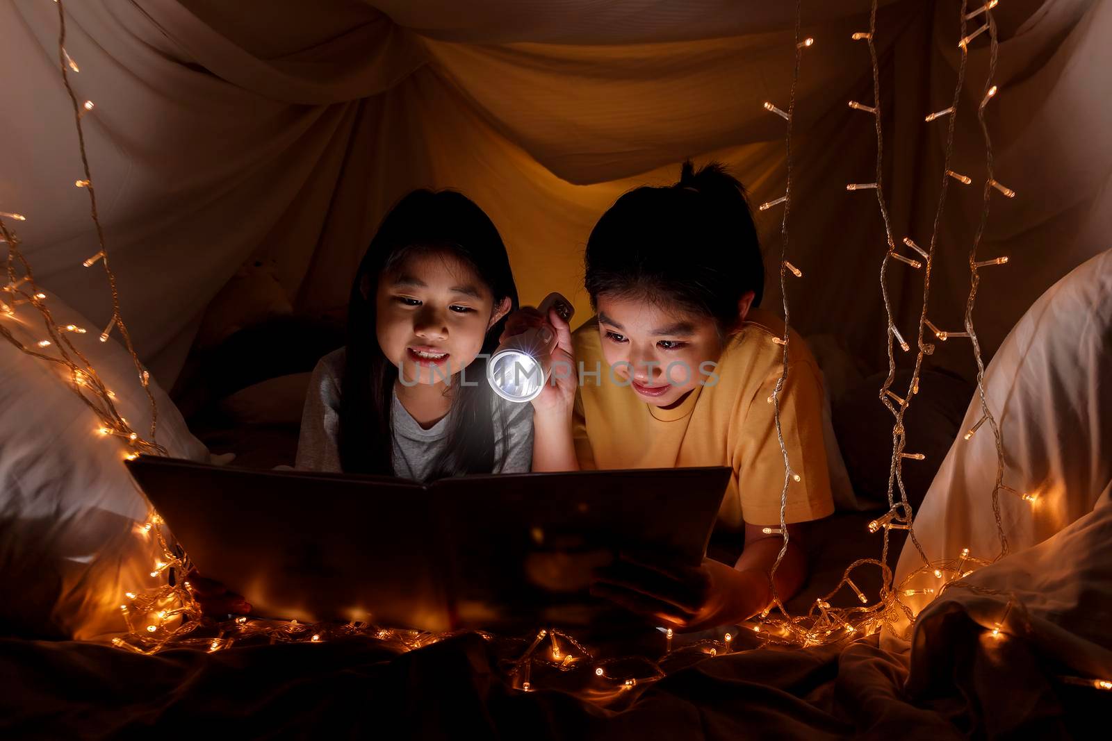 Family concept. elder sister and sister reading book with flashlight together before bedtime. Sister read story book together in bed sheet tent. focus selective little sister. With film grain effect by Satrinekarn