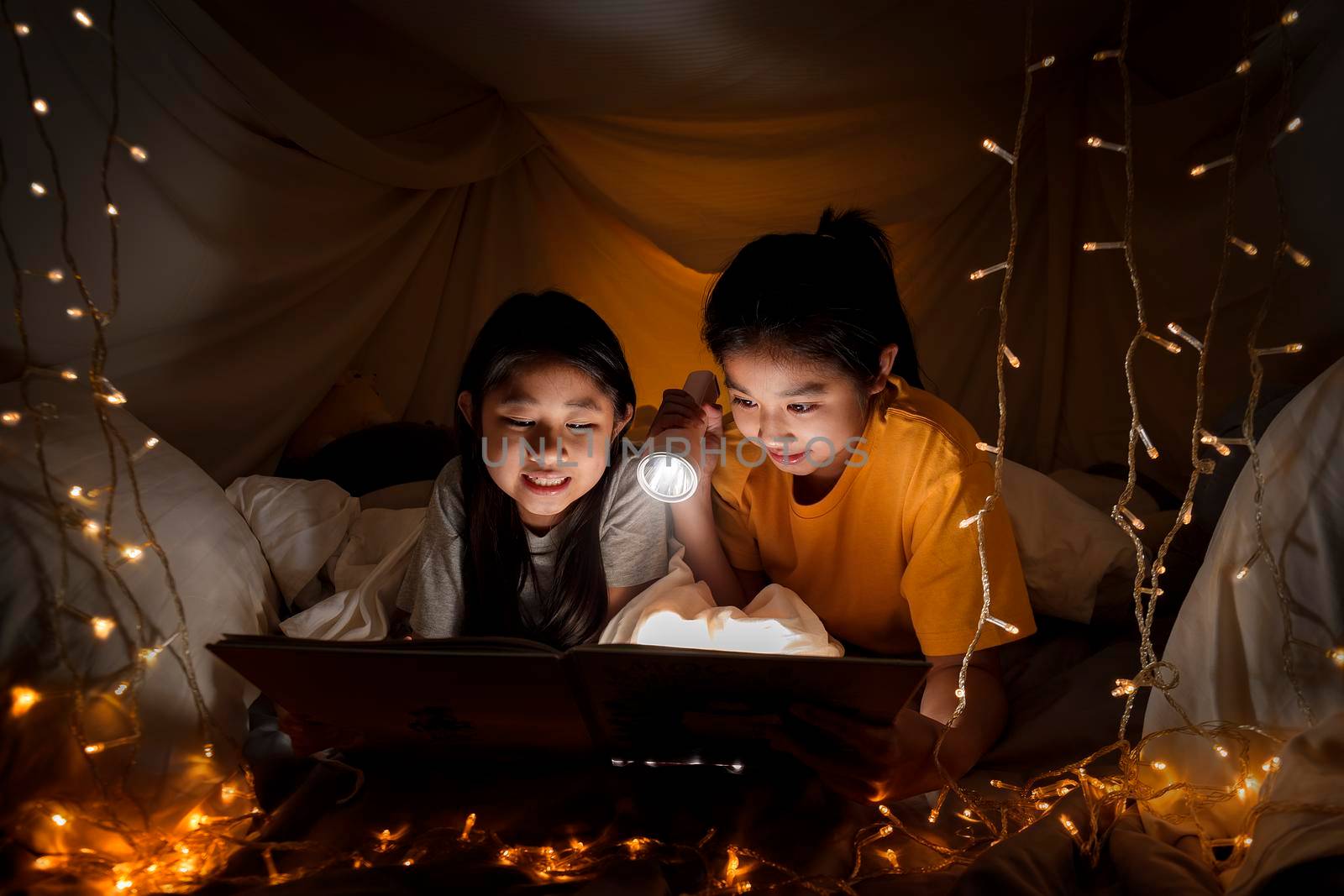 Family concept. elder sister and sister reading book with flashlight together before bedtime. Sister read story book together in bed sheet tent. focus selective little sister. With film grain effect by Satrinekarn