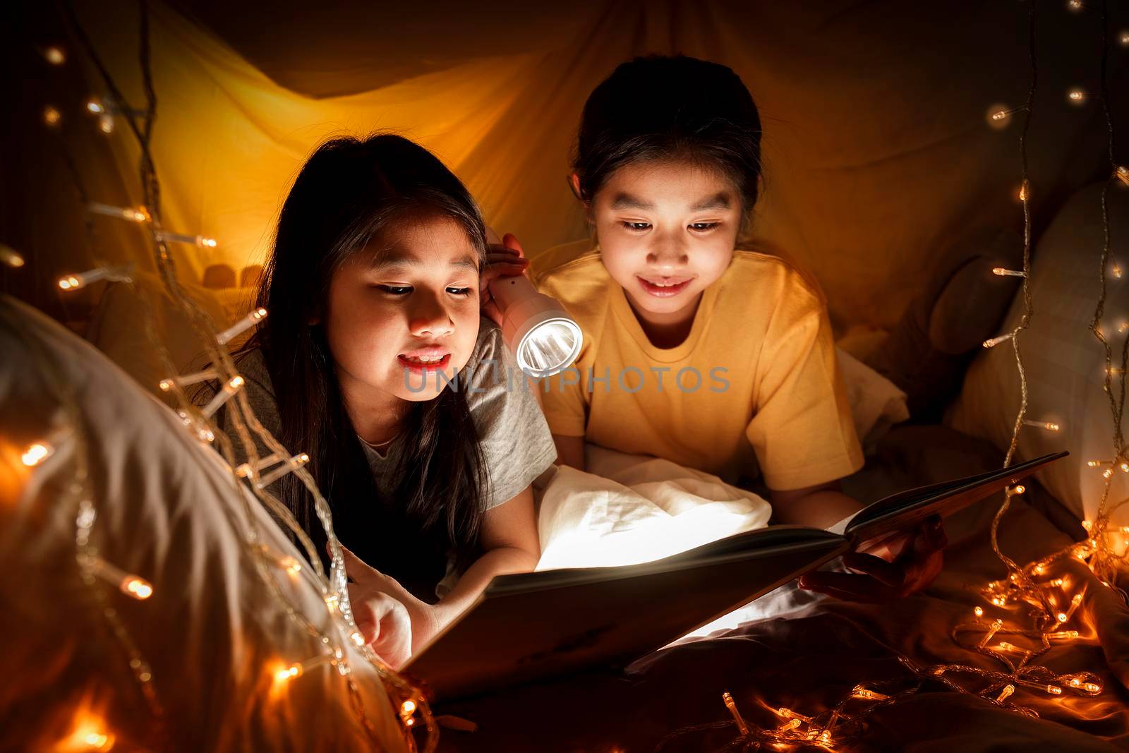 Family concept. elder sister and sister reading book with flashlight together before bedtime. Sister read story book together in bed sheet tent. focus selective little sister. With film grain effect by Satrinekarn