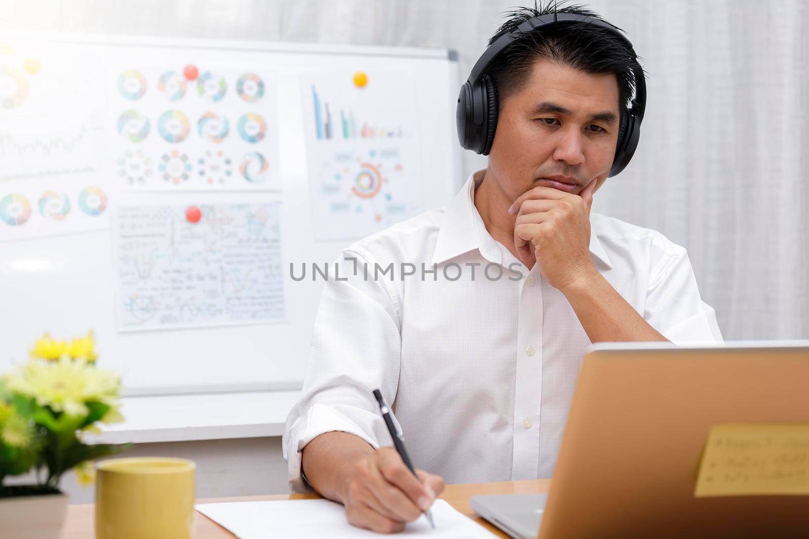 Asian businessman talking with team via video conference writing note on paper. business people using laptop and headset for online meeting. Man using headphone speak with customers. work from home concept.