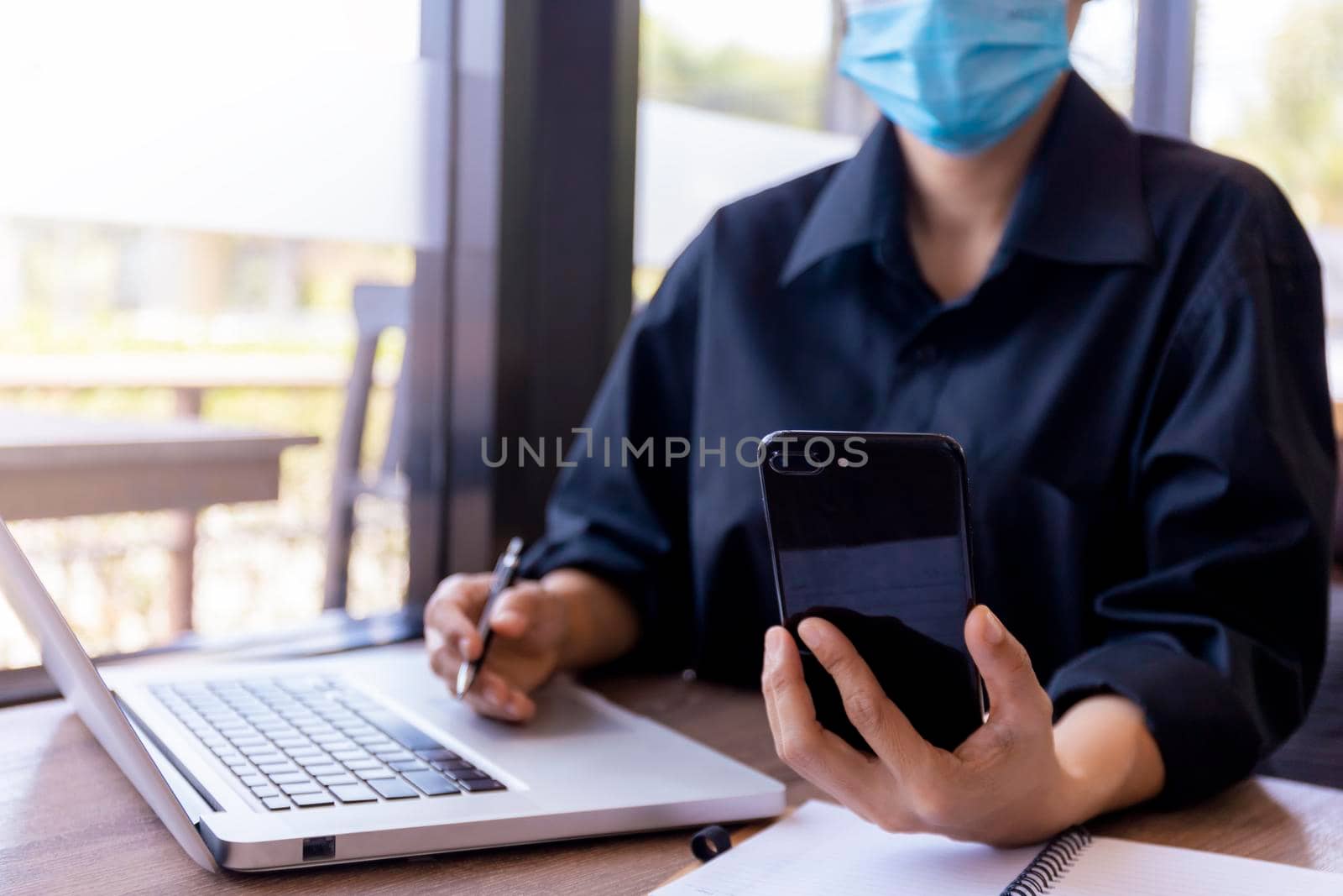 Young woman with protective face mask sitting in coffee shop at wood table using smartphone.and laptop. Girl browsing internet, chatting, blogging. Female holding phone and looking on screen. covid-19 new normal