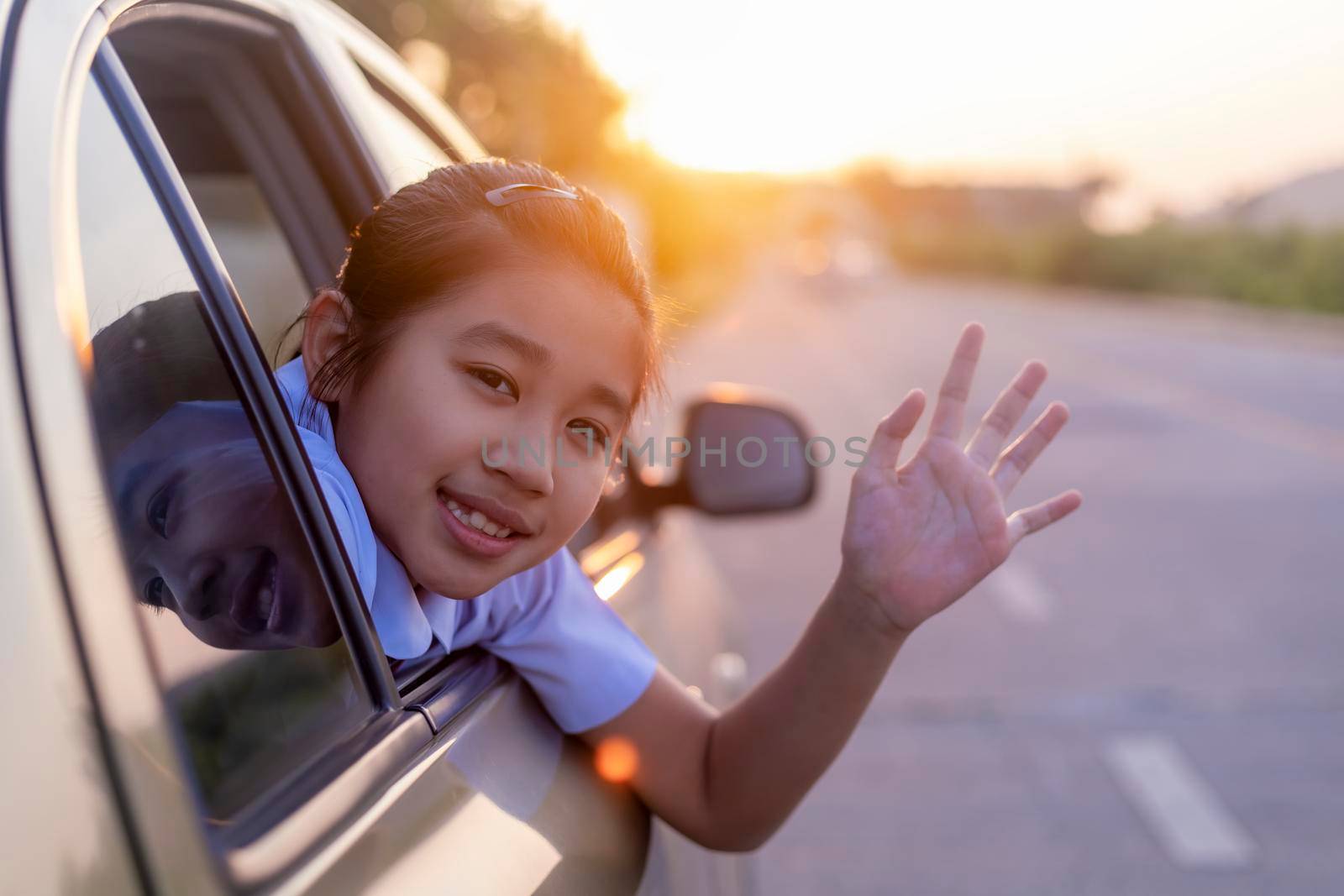 On the way to school, the little girl stretched out his hand from the car window, laughing and smiling. Asian little girl smiling  and waving hand out the car. Children relax with street view from the car. Family in car concept. by Satrinekarn