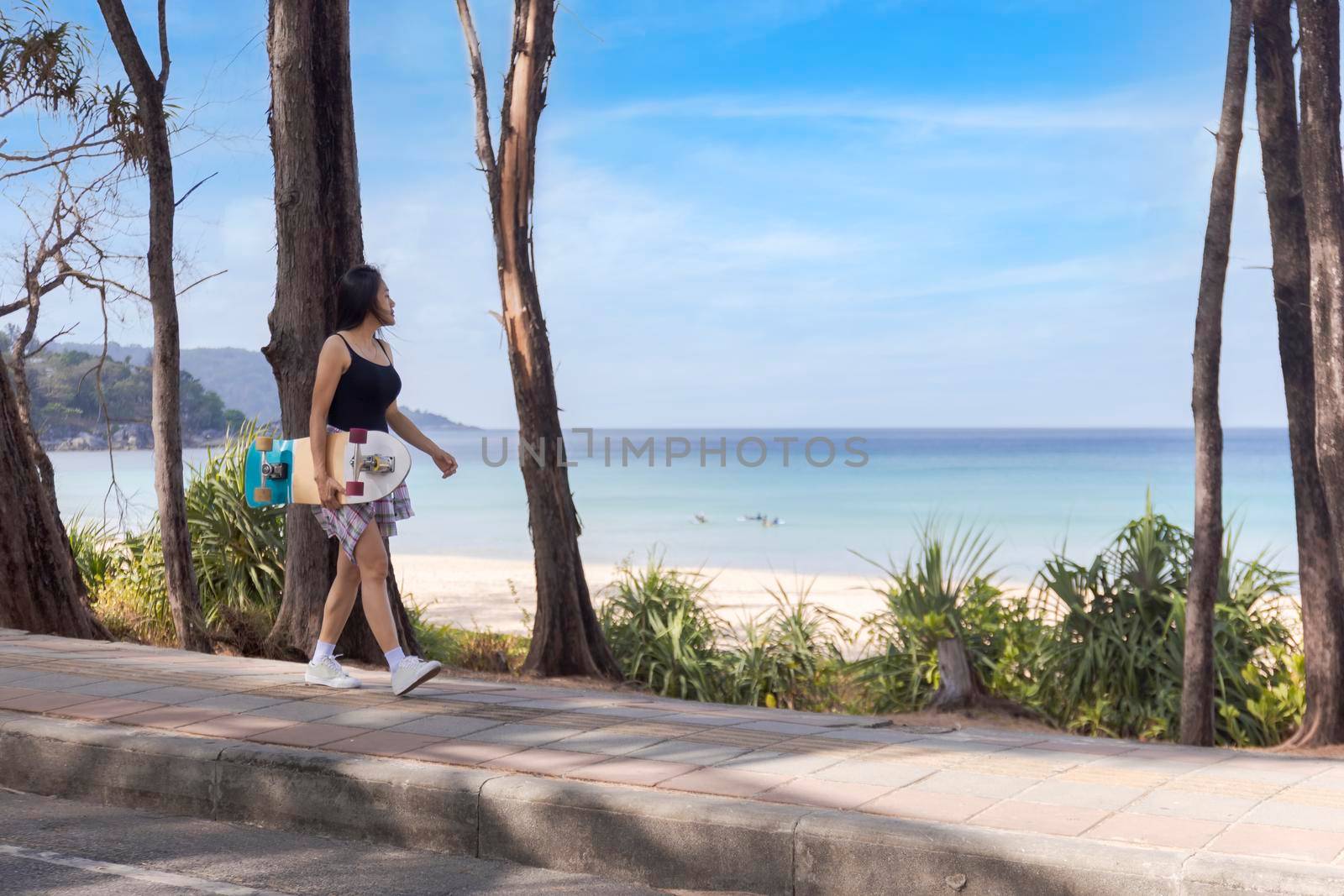 Young women walking on side road near tropical beach with surf skate board. Young girl long hair wear สong sleeve shirt, shot jeans and sneaker walk with surf skate board on the beach road. by Satrinekarn