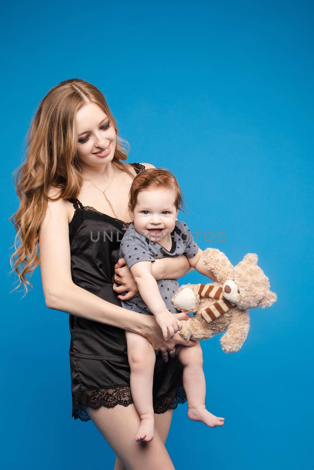 Front view of positive little baby laughing with mother in studio. Beautiful woman in black pajamas keeping cute daughter on hands. looking at camera and posing on blue isolated background.