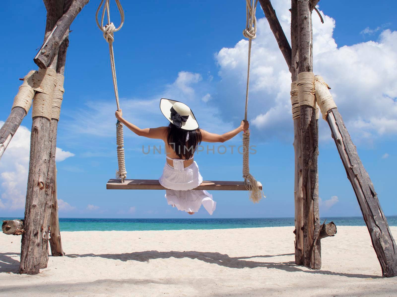 Happy young woman enjoy and sway the wooden swing over the sea beach, blue sky and cloud. Tourist women relax and enjoy with wood swing on the beach Phuket, Thailand. Summer holiday vacition concept.