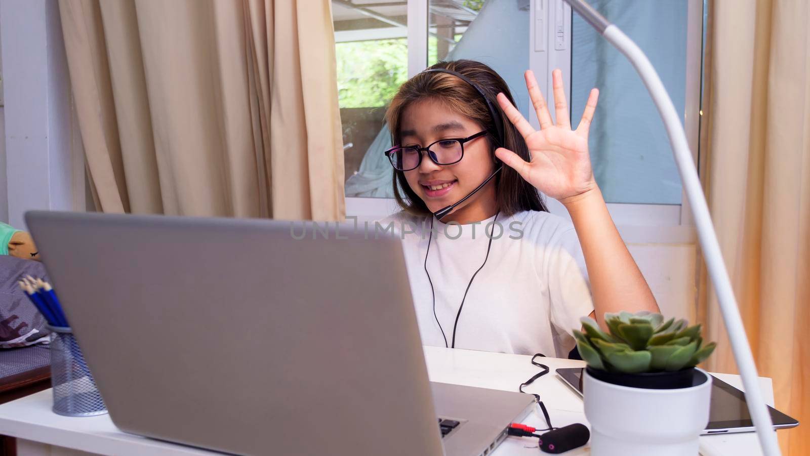  Asian girl in headphones sit at desk study online on laptop. Kid wear headset handwriting in notebook learning using internet lessons on quarantine. Girl student learning virtual internet online class from school due to covid pandemic.