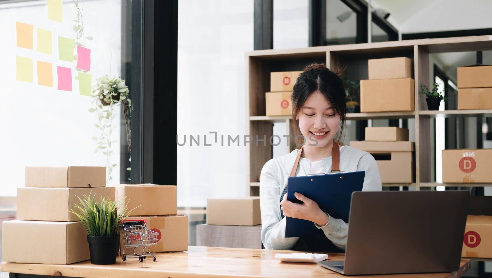 A portrait of a young Asian woman, e-commerce employee sitting in the office full of packages in the background write note of orders and a calculator, for SME business, e-commerce and delivery business. by wichayada