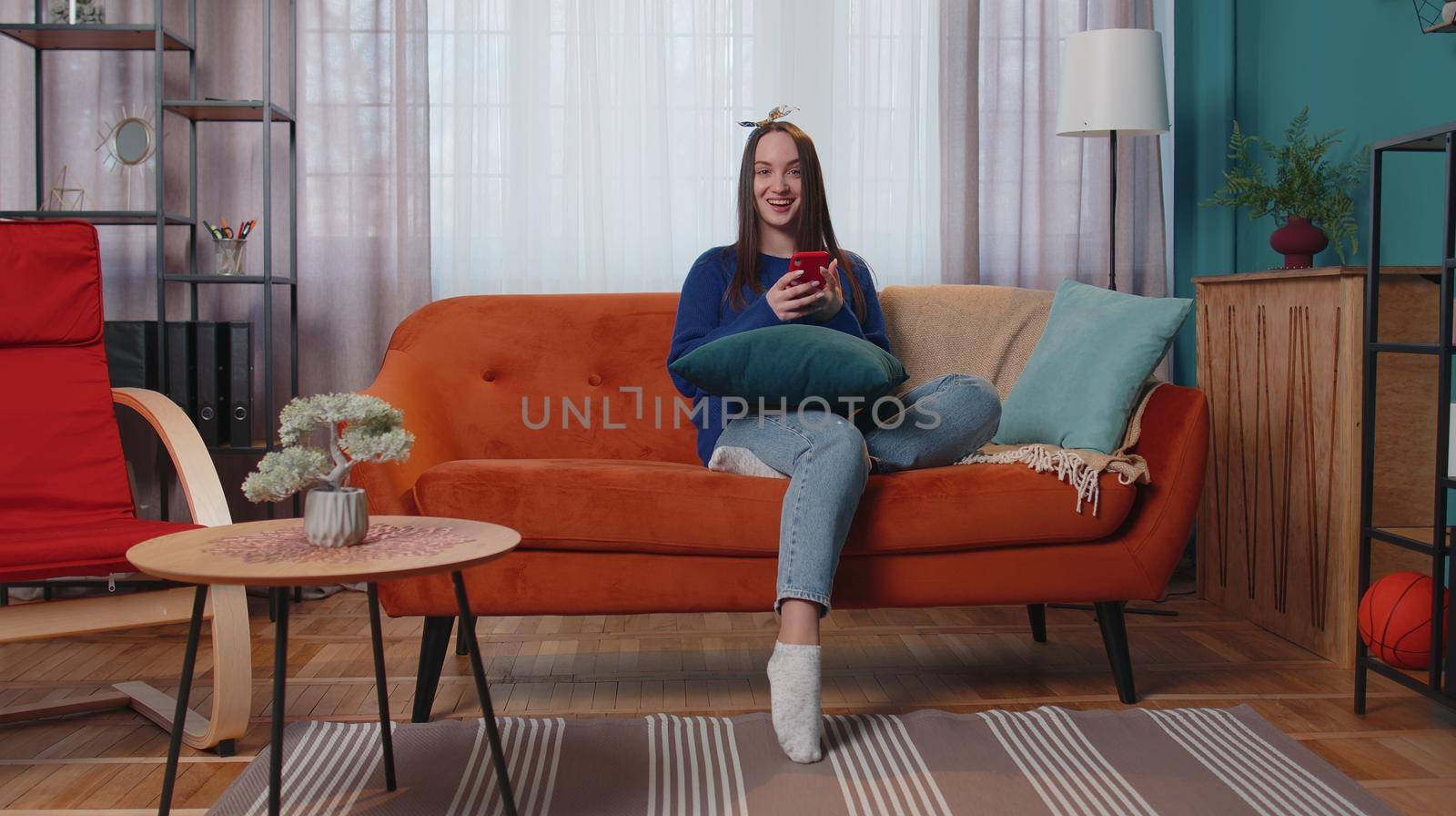 Happy excited joyful stylish teen girl in sweater use mobile cell phone typing browsing say wow yes found out great big win news doing winner gesture. Young adult woman at home in living room on sofa