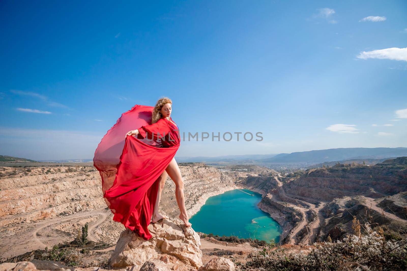 Side view of a beautiful sensual woman in a red long dress posing on a rock high above the lake in the afternoon. Against the background of the blue sky and the lake in the form of a heart.