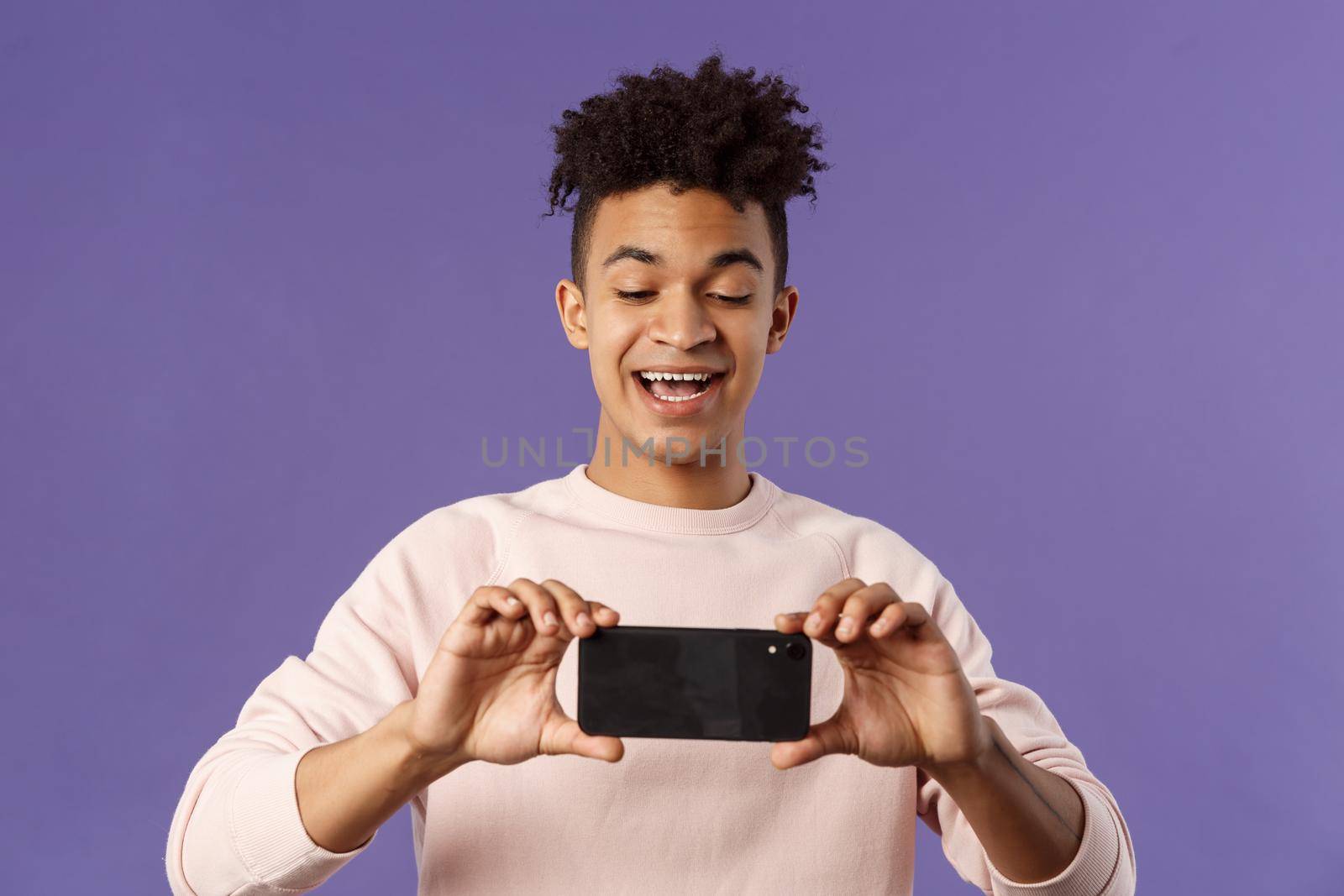 Portrait of amazed, excited young man seeing something interesting, stream concert to his internet social network profile, taking photo or recording video with mobile phone, purple background.
